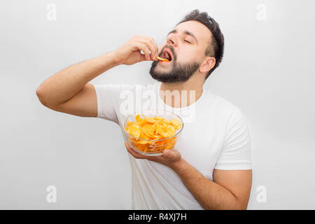 Man in white shirt stands and eats chips. He puts it in mouth with hand. Also guy has a bowl with them in another hand. It's delicious. Isolated on white background. Stock Photo