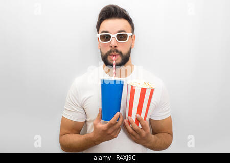 Confident young man looks on camera through movie glasses. He holds glass of coke nad popcorn in both hands. Young man drinks through straw. Isolated on white background. Stock Photo