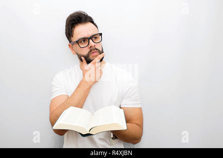Wize and thoughtful young man in glasses stand and hold opened book in hands. He looks to side. Guy holds hand on chin. Isolated on white background. Stock Photo