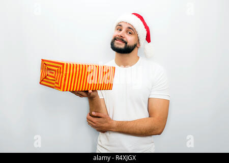 Nice bearded man in red hat stands and holds orange box with present in one hand. He looks on camera and smiles a bit Isolated on white background. Stock Photo
