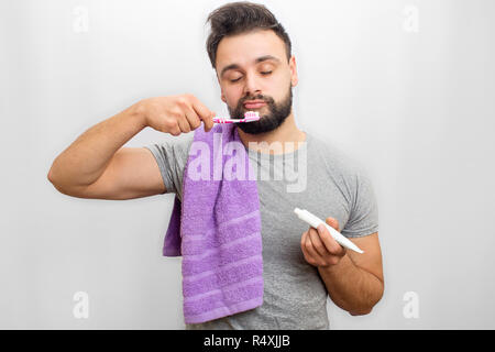 Oicture of sleepy young man holding tooth brush in one hand and tooth paste in another one. He keeps eyes closed. Also guy has towel across shoulder. Isolated on white background. Stock Photo