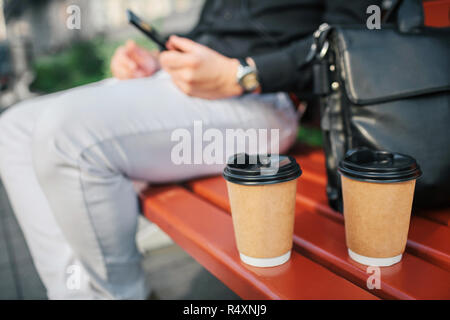 Cut vuew of man sitting on bench. There are cup nd black leather bag. He holds phone in hands. Guy sit outside. Stock Photo