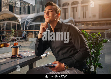 Thoughtful young man sit at table outside. He looks up. It is sunny. Stock Photo