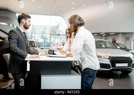 Young business couple with salesperson or manager buying or renting car standing at the stand in the showroom Stock Photo