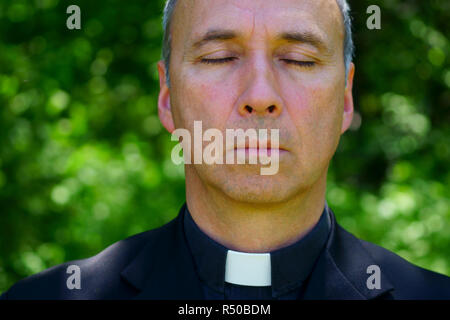 A good looking catholic priest is praying in the forest. Eyes closed, concentrating. Stock Photo