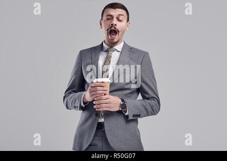 Portrait of handsome young confident arabic businessman with fancy mustache in fashion gray full suit yawns with a cup of coffee in his hands on studi Stock Photo