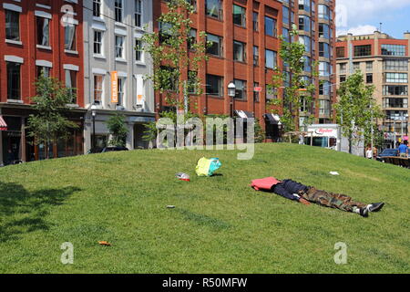 Possibly homeless man lays on grass with a shirt covering his head, surrounded by litter. Stock Photo