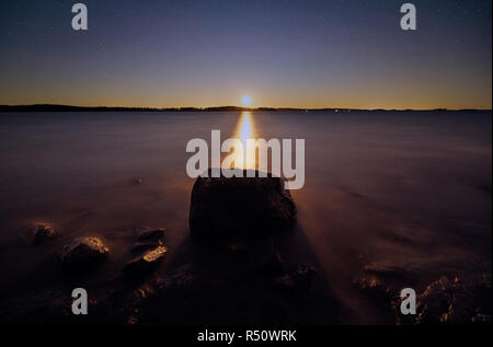 Moon shining just above the horizon at night. Big stone on still water of peaceful lake in Finland. Stock Photo