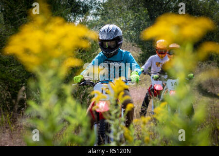 Two boys and a man riding motorcycles in forest behind plants Stock Photo