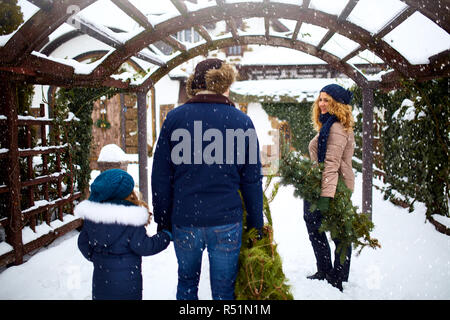 Authentic family carrying Christmas trees and branches to house. Father mother and daughter prepare for New Year and going to decorate their home with spruce and fir tree on celebration. Snowfall. Stock Photo