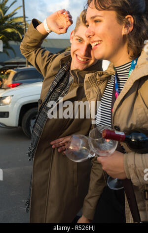 Young boy and girl or couple between 18 to 22 years of age walking in the street carrying a bottle of red wine and two empty glasses smiling and happy Stock Photo