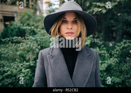 Photo of young blonde in gray coat on blurred background of city, green trees Stock Photo