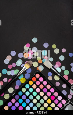 Cosmetic background with makeup brushes and eyeshadow palette on a black background with scattered confetti Stock Photo