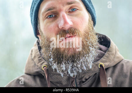 Portrait of a looking at camera young man in a gray hat and a brown jacket with a long beard covered with icicles. Men winter joys concept Stock Photo