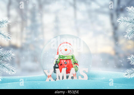 Cute snowman in Santa Claus clothes in New Year, Christmas glass magic ball. Snow fall on it and Christmas tree beside. XMAS letters beside. Free spac Stock Photo