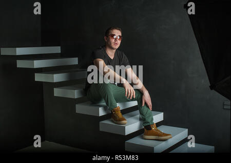 studio photography young brutal guy. man in sunglasses, T-shirt, jeans and high boots on a background of black wall on the white steps. sitting on the steps, protinuv hands forward, putting them on her knees Stock Photo