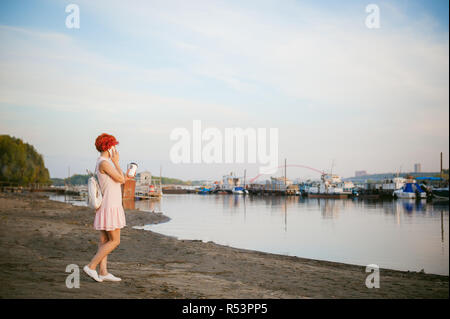 girl in pale pink dress with red hair and backpack walking along river bank, talking on the phone and drinking coffee from a cardboard cup, against backdrop of boats moored on a warm summer day Stock Photo