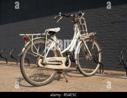 Old rusty grunge vintage white bike in bicycle rack in front of a black painted brick wall. Stock Photo
