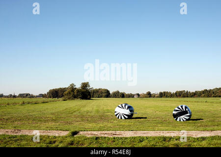 Two bales of hay wrapped in black and white striped plastic and stacked in a landscape with trees at the horizon. Stock Photo