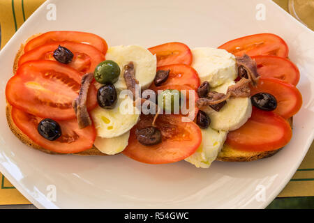 Lunchtime snack of tomatoes, cheese. anchovies and olives on bread, served on a white plate. Stock Photo