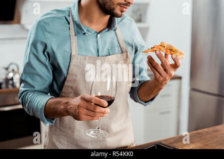 cropped shot of man in apron holding glass of wine and slice of homemade pizza Stock Photo