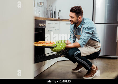 smiling young man in apron taking out baking tray with pizza from oven Stock Photo