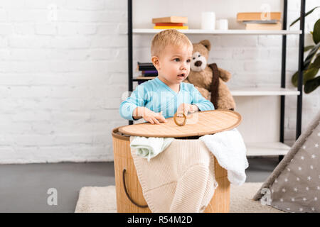 cute baby in blue bodysuit standing near wicker basket with laundry in light room Stock Photo