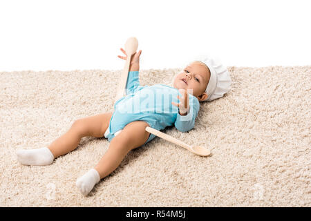 toddler boy in chefs hat with two big wooden spoons lying on carpet and looking at camera isolated on white Stock Photo