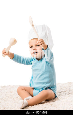 toddler boy in chefs hat sitting on carpet and holding two big wooden spoons isolated on white Stock Photo