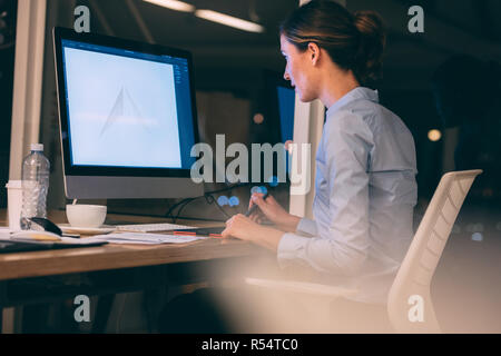 Woman doing graphic designing sitting at her desk in office late in the night. Woman using a digital writing pad to make designs on computer. Stock Photo