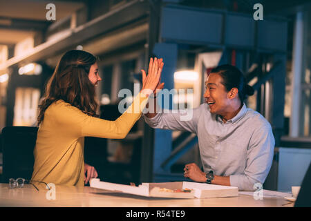 Smiling business colleagues sitting late in office celebrating project completion. Happy businessman and woman eating pizza sitting late in office aft Stock Photo