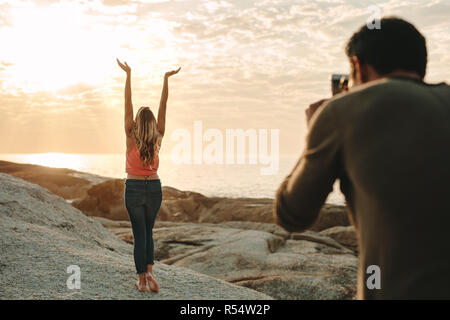 Man taking photo of a woman standing on a rock at the beach using a mobile phone. Rear view of woman with her arms raised and looking up with sun and Stock Photo