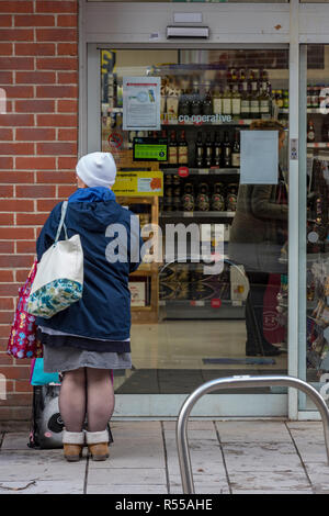 an old lady carrying a shopping bag standing outside of a supermarket looking in the window of the shop. Stock Photo