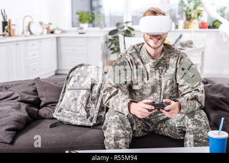 smiling army soldier in virtual reality headset playing video game on couch Stock Photo