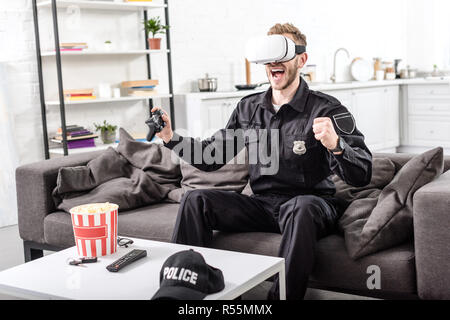 policeman with virtual reality headset on head playing video game and screaming with joy on couch Stock Photo