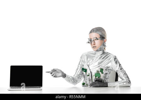 silver robot sitting at table and pointing on laptop with blank screen isolated on white, future technology concept Stock Photo