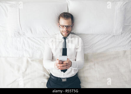Top view. Handsome businessman with glasses lying on bed texting from his smartphone Stock Photo