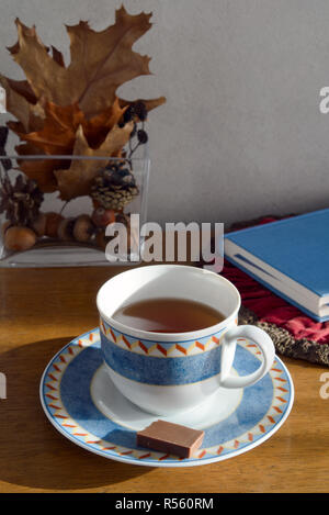 still life with cup of tea and a piece of chocolate, in the background a book and decoration of autumn dry leaves (cup with saucer separated) Stock Photo