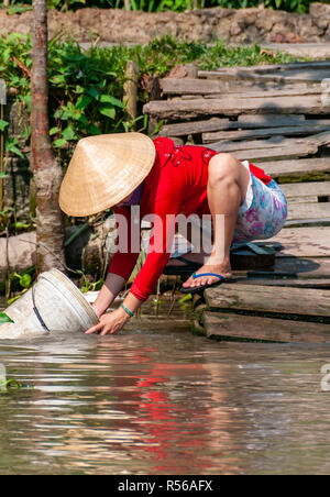 Young woman wearing bright red top crouching by the water's edge to wash out a bucket in river at Can Tho Province, Vietnam Stock Photo