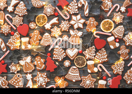 Christmas pattern made of gingerbread cookies, anise stars, allspices, berries, candy cane lollipops, baking molds, orange chips and wooden decorations with garland on wooden background. Flat lay. Stock Photo
