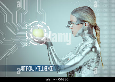side view of cyborg holding and examining green apple with digital data isolated on grey with 'artificial intelligence' lettering Stock Photo