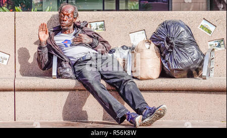 An African-American homeless senior citizen male sits on a park bench in Pershing Square in Los Angeles and gestures with his right hand, holding his  Stock Photo