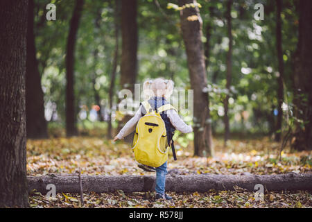 The theme children outdoor activities. Funny little baby Caucasian blond girl walks through forest overcoming obstacles, tree fell, log. Baby hiking big funny backpack in autumn forest park Stock Photo