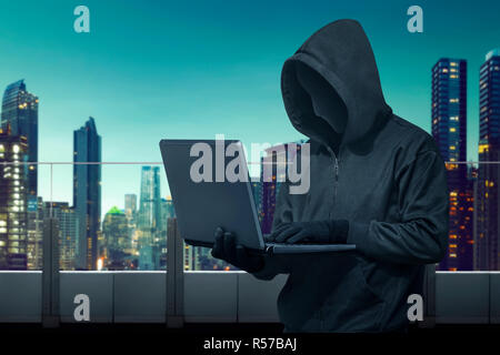 Hooded hacker with vendetta mask stealing information with laptop Stock Photo