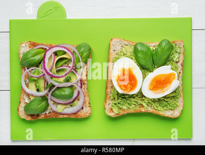Two avocado toasts with soft boiled eggs,mashed avocado,sliced avocado ,red onion,basil leaves and goat cheese on green background Stock Photo