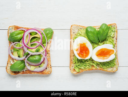 Two avocado toasts with soft boiled eggs,mashed avocado,sliced avocado ,red onion,basil leaves and goat cheese on white wooden background Stock Photo