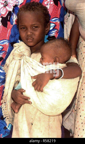 1993 - Straight on, medium close-up of a young Somali girl cradling her sister in sling that is tied over her shoulder.  They are waiting in line to see US Navy doctors conducting a medical civic action program in the capital city of Mogadishu, Somalia.