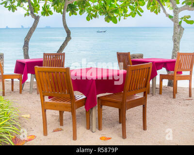 Wooden dinner table set with red tablecloth on the sand beach with the long-tail boat and seascape background. Stock Photo