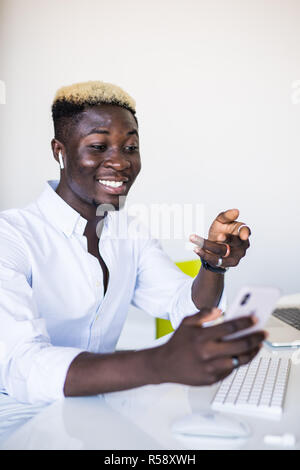 Young African American man employee working on generic laptop computer and looking at phone with smile in office Stock Photo