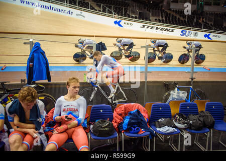 Berlin, Germany. 30th Nov, 2018. Cycling: Track Bike World Cup. A cyclist of the national team of Russia keeps himself warm at the edge of the qualifications of team pursuit in the velodrome with a training bike. In the background the national team of Germany passes by during the men's qualification. Credit: Gregor Fischer/dpa/Alamy Live News Stock Photo